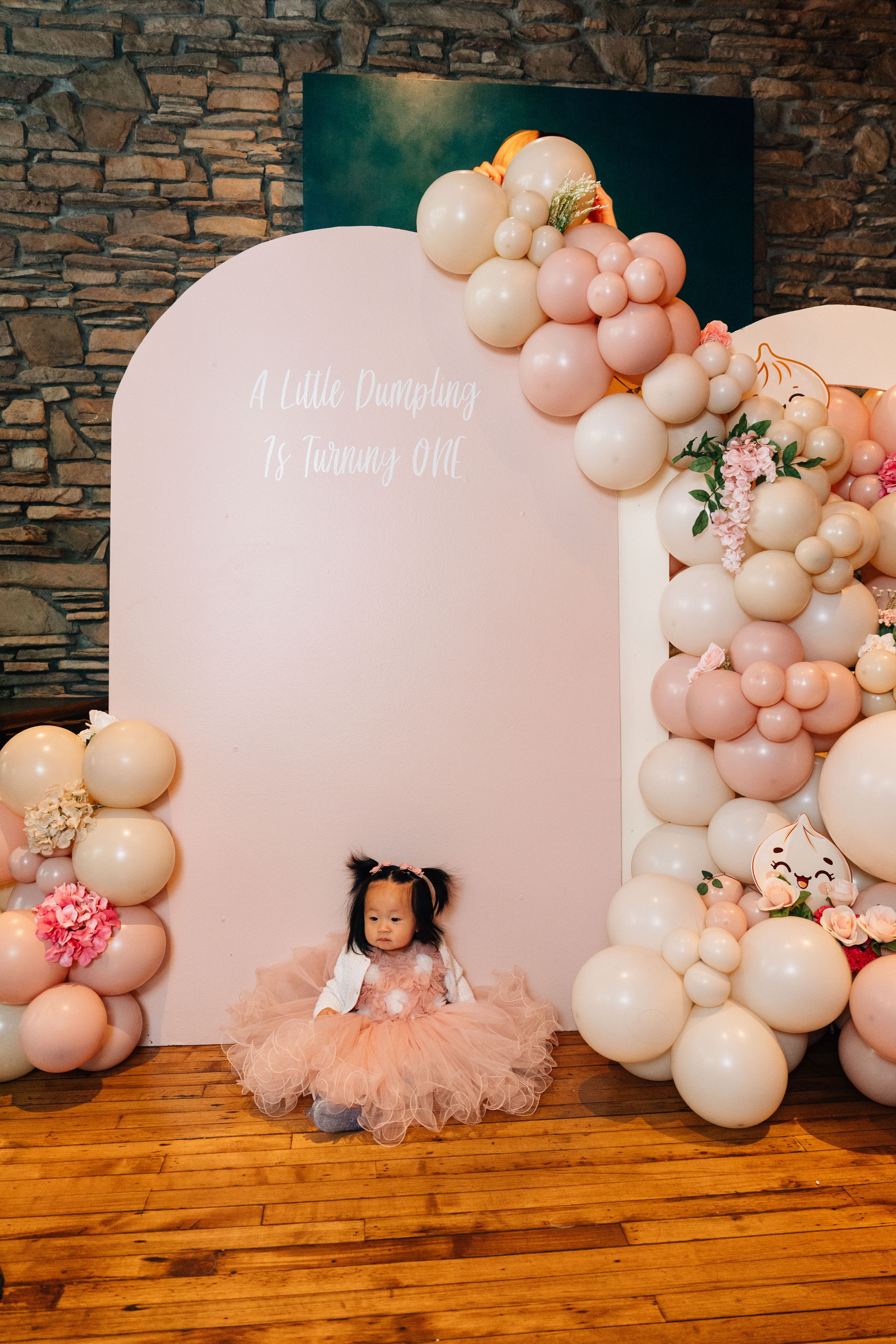 Pink and white balloon arch decorates a birthday party backdrop with floral accents and hardwood flooring.