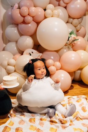 A baby lays on a white blanket with peach-colored balloon decorations and floral accents for a milestone photoshoot.