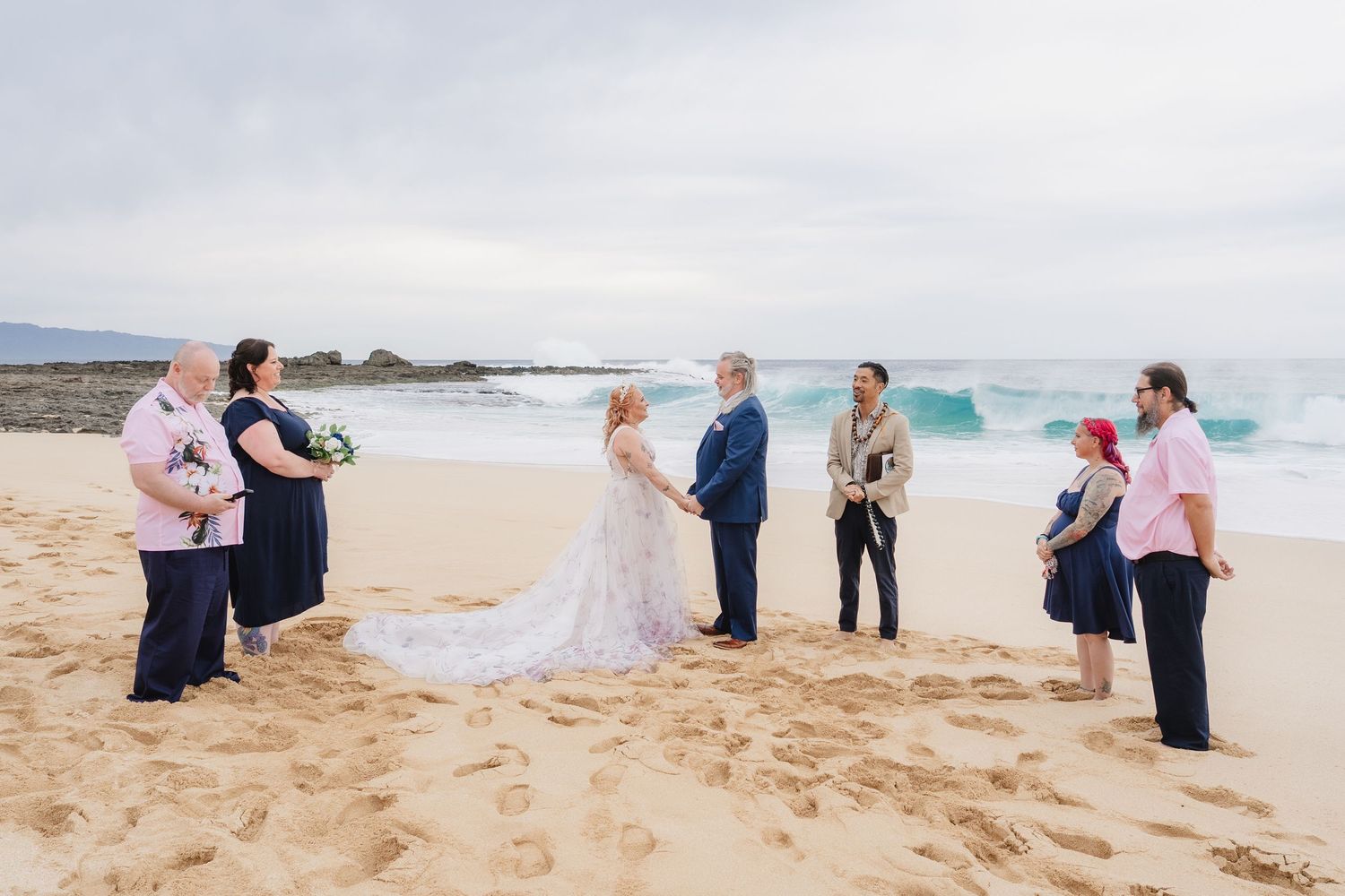 A group gathers on a sandy beach as waves crash behind them during an outdoor celebration.