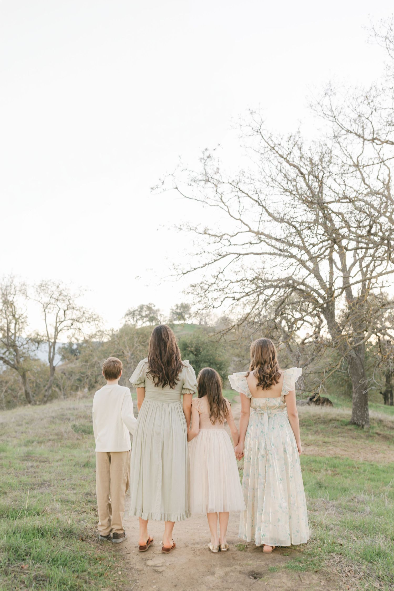 A group of four people wearing light colored dresses and clothing stand together on a grassy hillside at sunset.