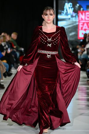 Burgundy velvet gown with flowing cape and silver embellishments showcased on runway at fashion show.