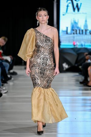 Model walks runway in metallic patterned cocktail dress with gold skirt during fashion show.