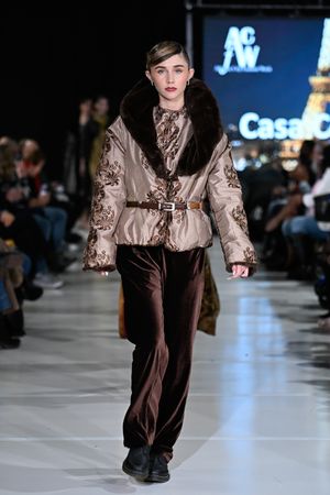 Fashion model walks runway in beige patterned winter coat with fur collar and brown pants at fashion show.