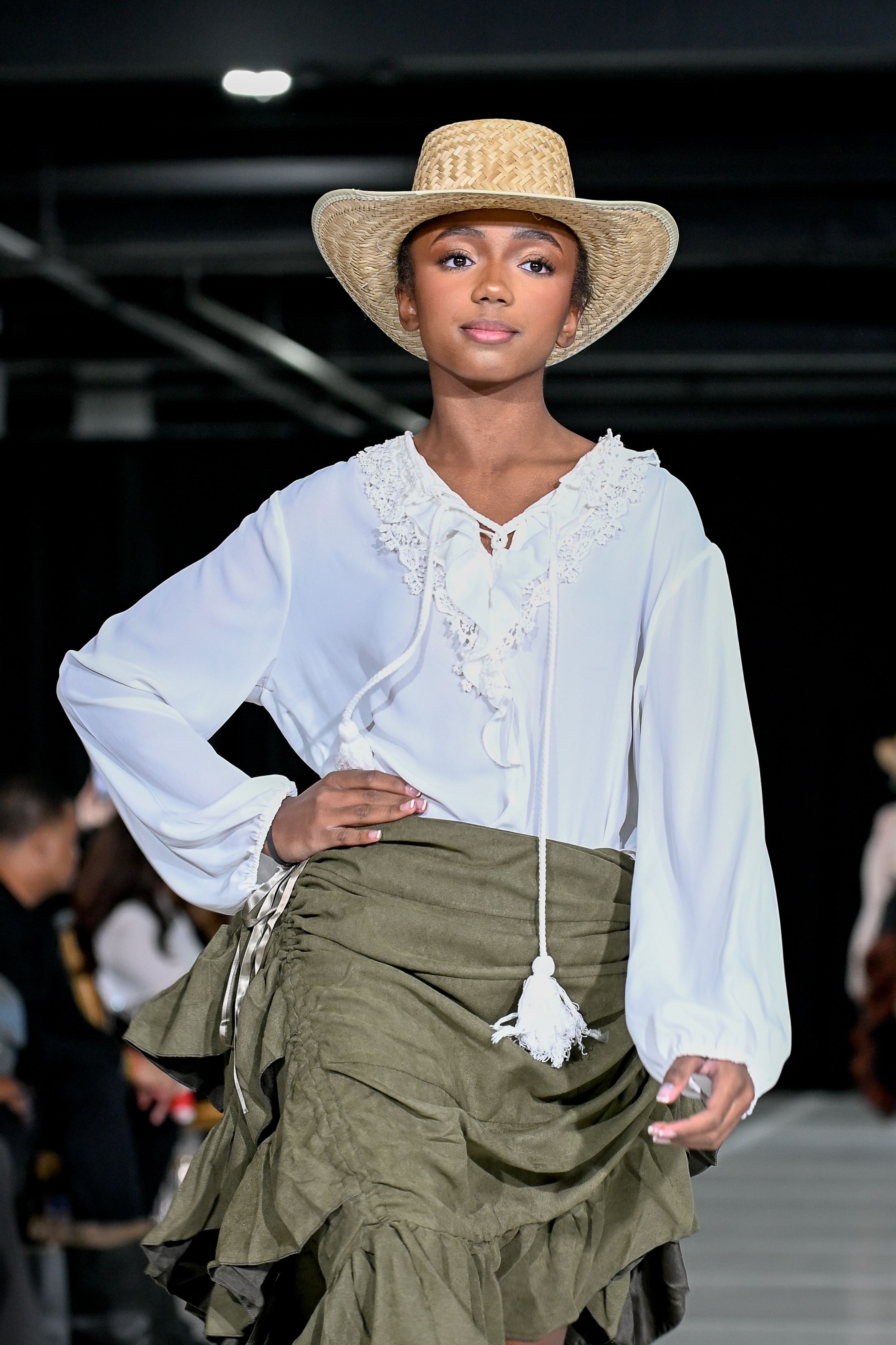 Model walks runway in white Victorian-style blouse and olive green skirt with straw hat at fashion show.