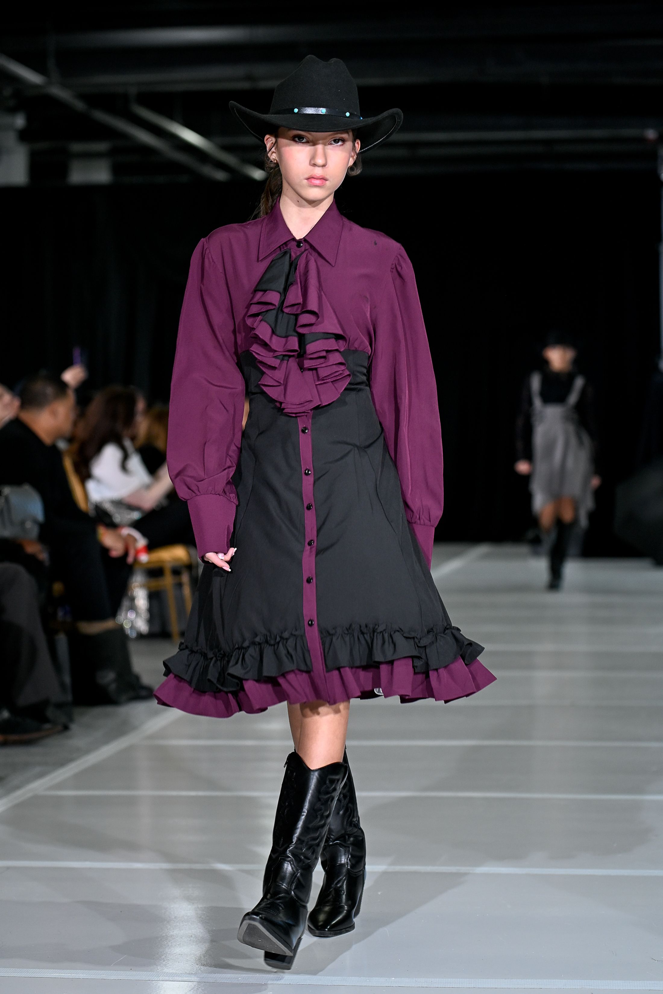 Fashion model walks runway wearing purple and black ruffled dress with black boots in dark industrial setting.