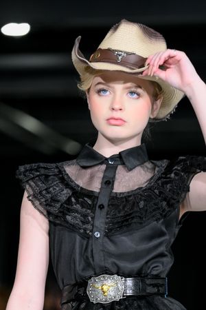 Fashion model wearing black lace top and brown fedora hat poses on dark runway.
