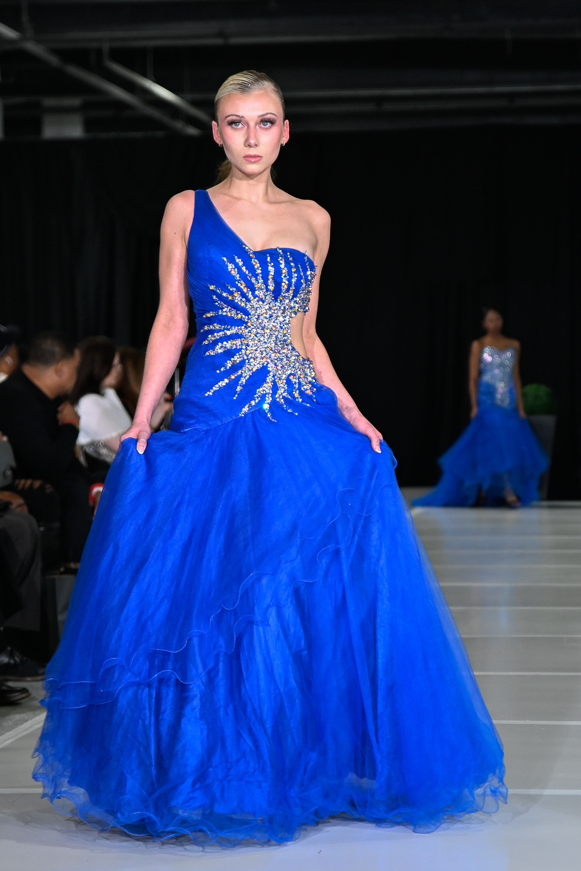 A model walks the runway in a vibrant royal blue tulle gown with crystal starburst detail at a fashion show.