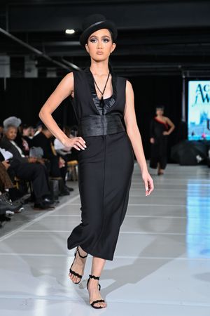 Model walks runway in black sleeveless dress and heels during fashion show in dark venue with blue lighting.
