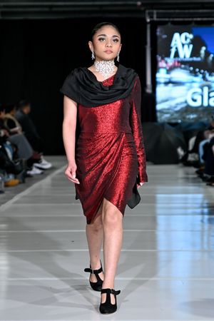 Model walks runway in red sequined cocktail dress with black shawl and heels at fashion show.