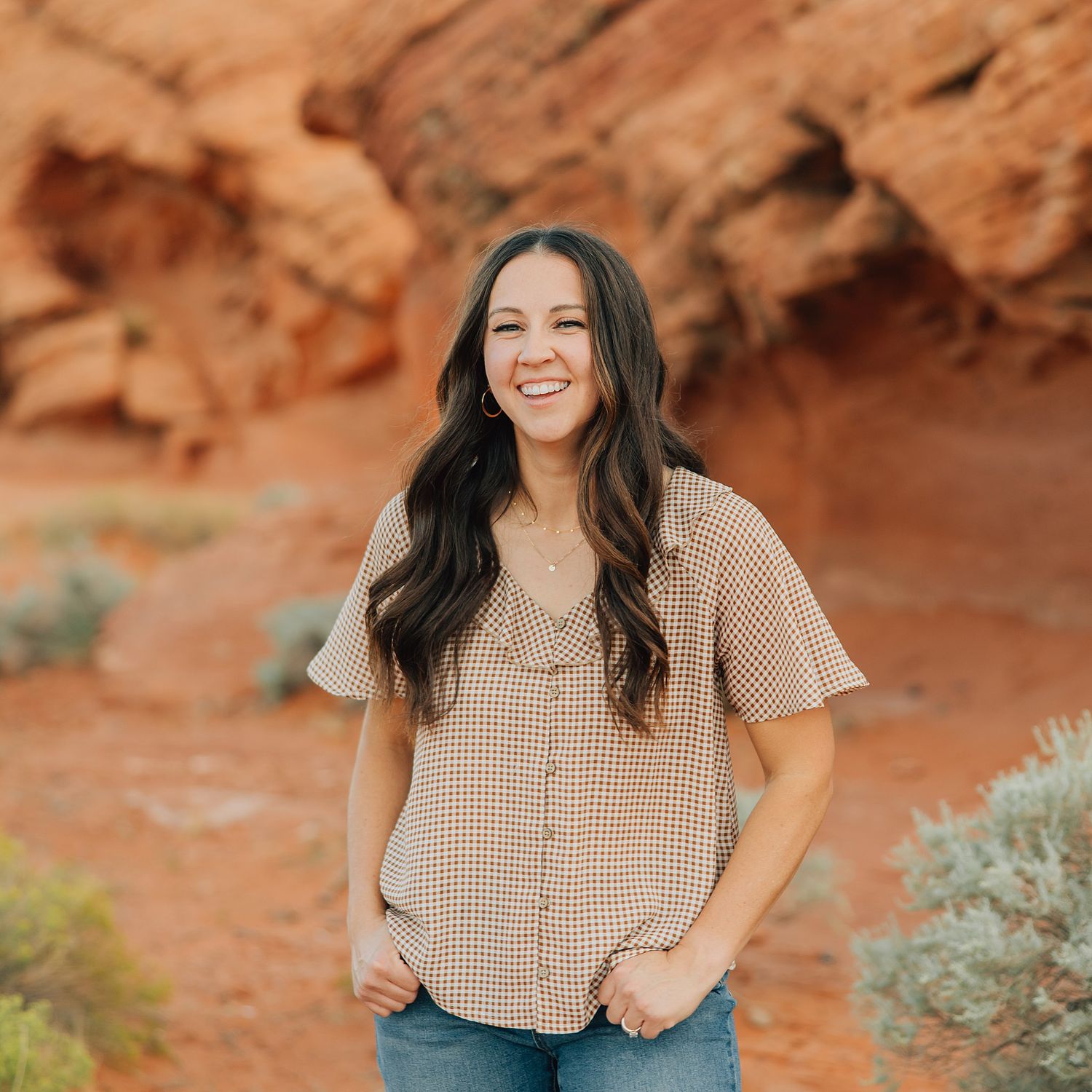 A person in a cream-colored dress poses against a desert landscape with red rocks and brush in the background.