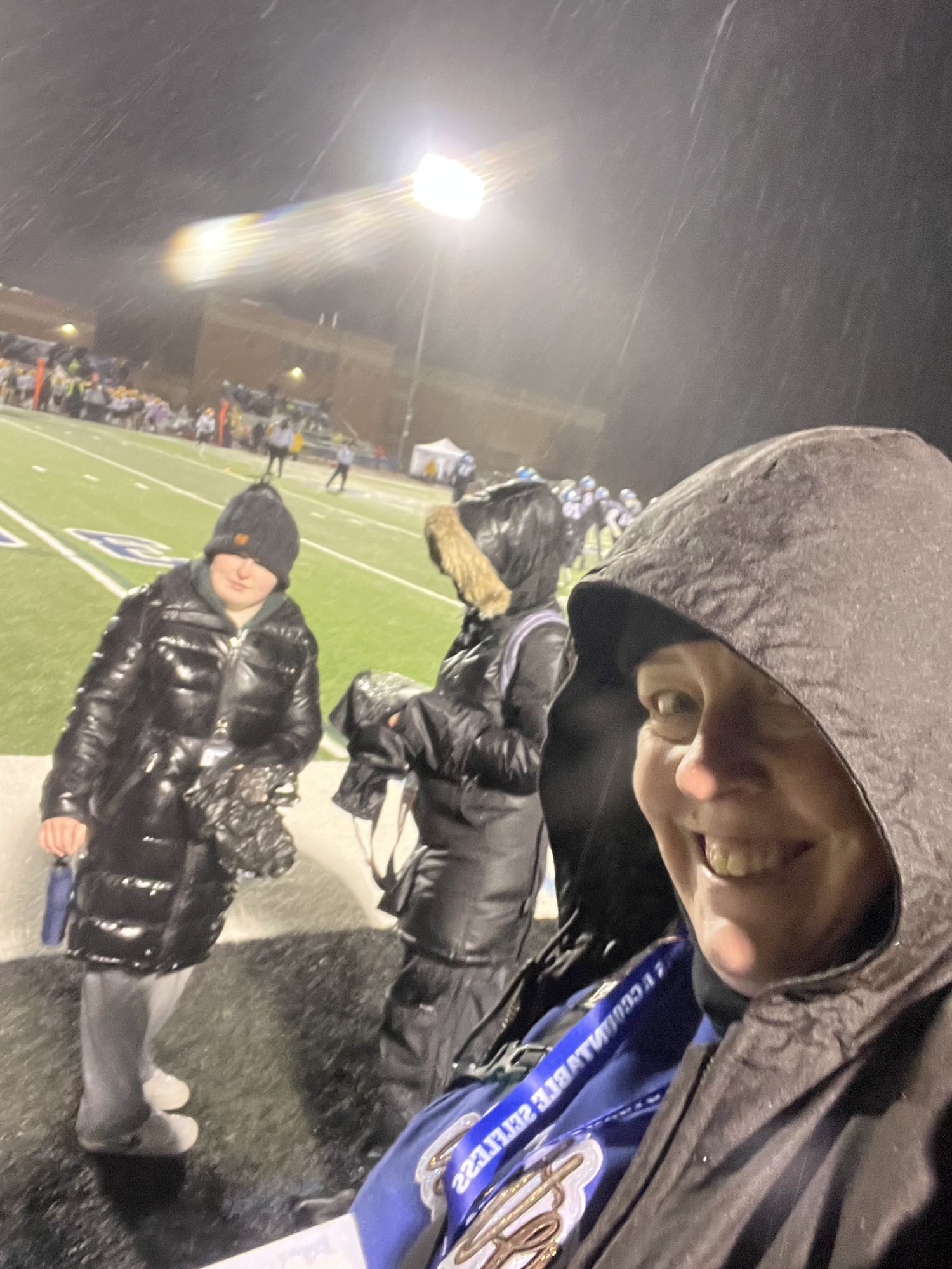 Photographers with cameras and winter gear working in snowy conditions at a nighttime football game on an illuminated field.