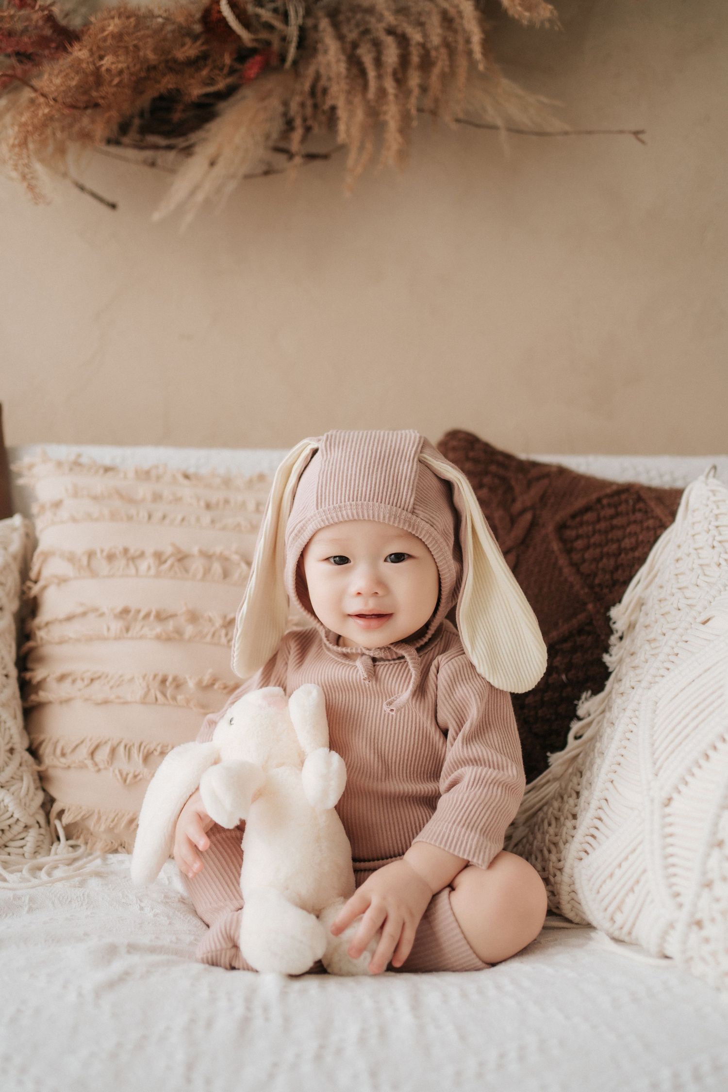 A baby wearing a cozy beige knit outfit lies on white bedding with plush bunny toys.
