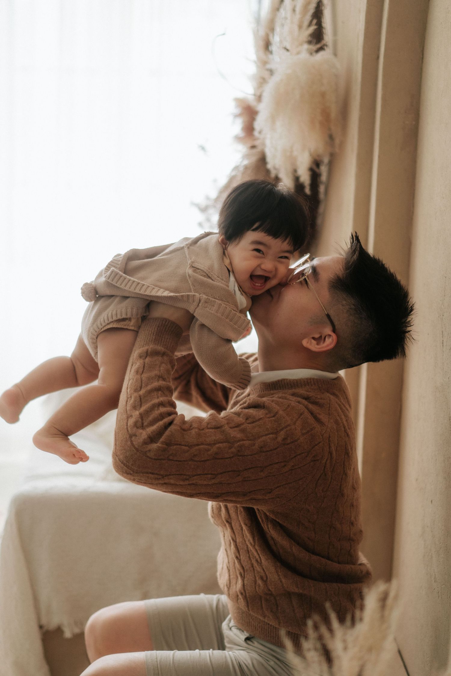 A man in a brown cable-knit sweater lifts a laughing baby in a cozy indoor setting.在溫馨的室內環境中，一名身穿棕色粗針毛衣的男子抱起一個咯咯笑的嬰兒。