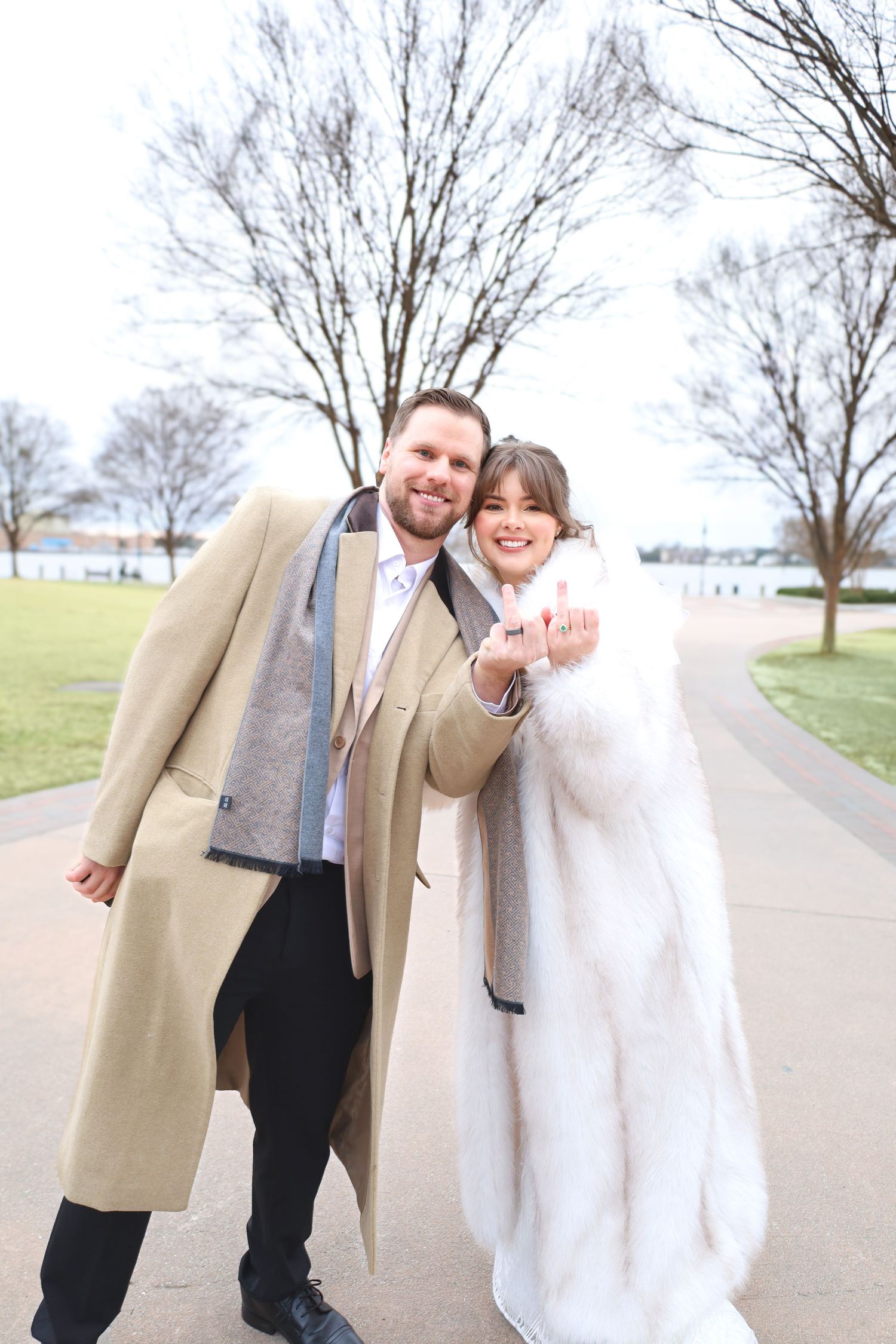 Couple in formal wear sharing a romantic moment outdoors on a paved path with bare trees in the background.
