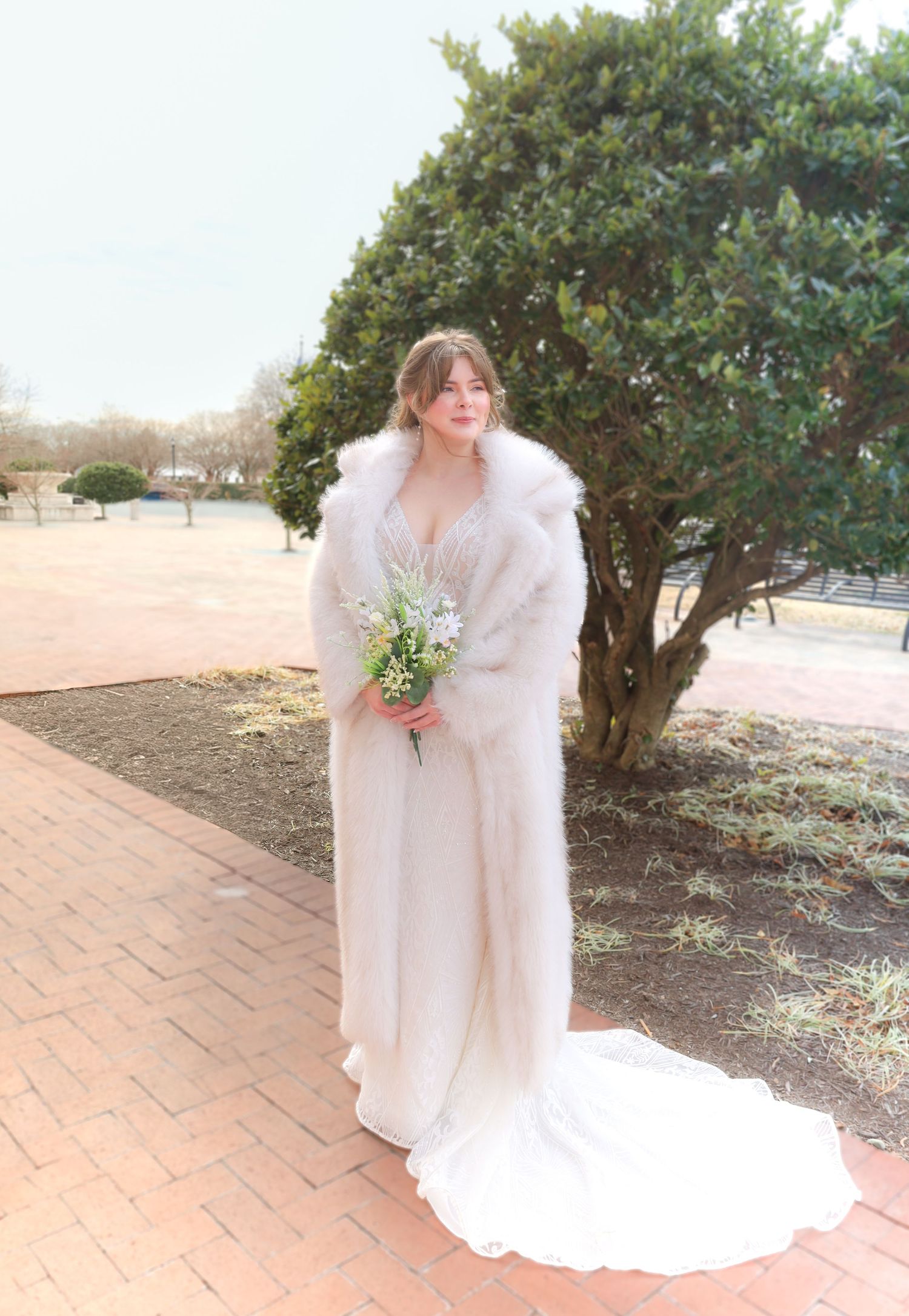 Winter bride in flowing white dress and fur stole holding delicate bouquet on brick path.