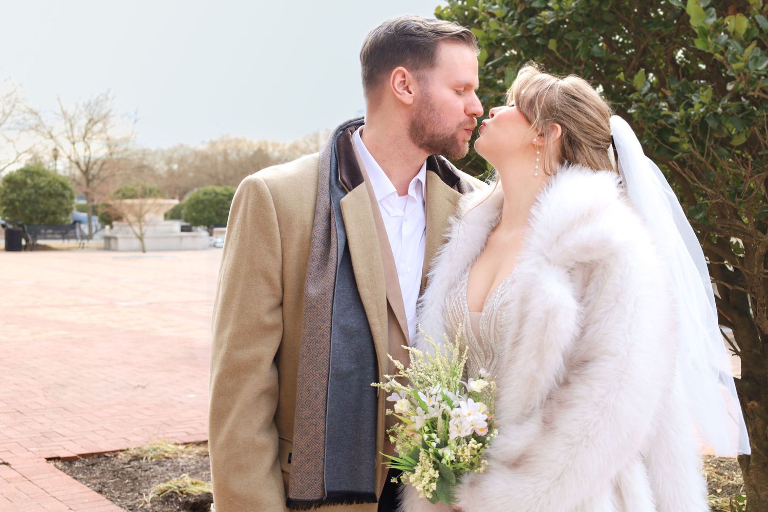 Couple embracing outdoors on a sunny day wearing formal attire near greenery.