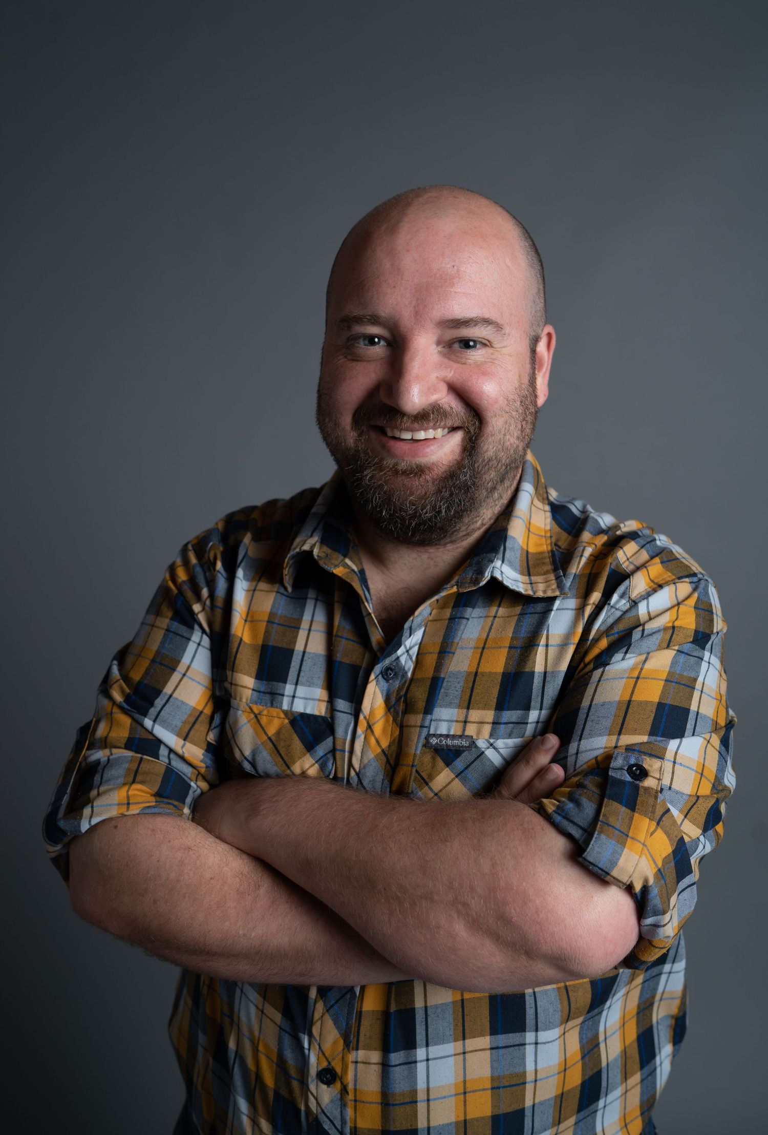 Portrait of a cheerful person in a yellow and blue plaid flannel shirt with crossed arms against a gray background.