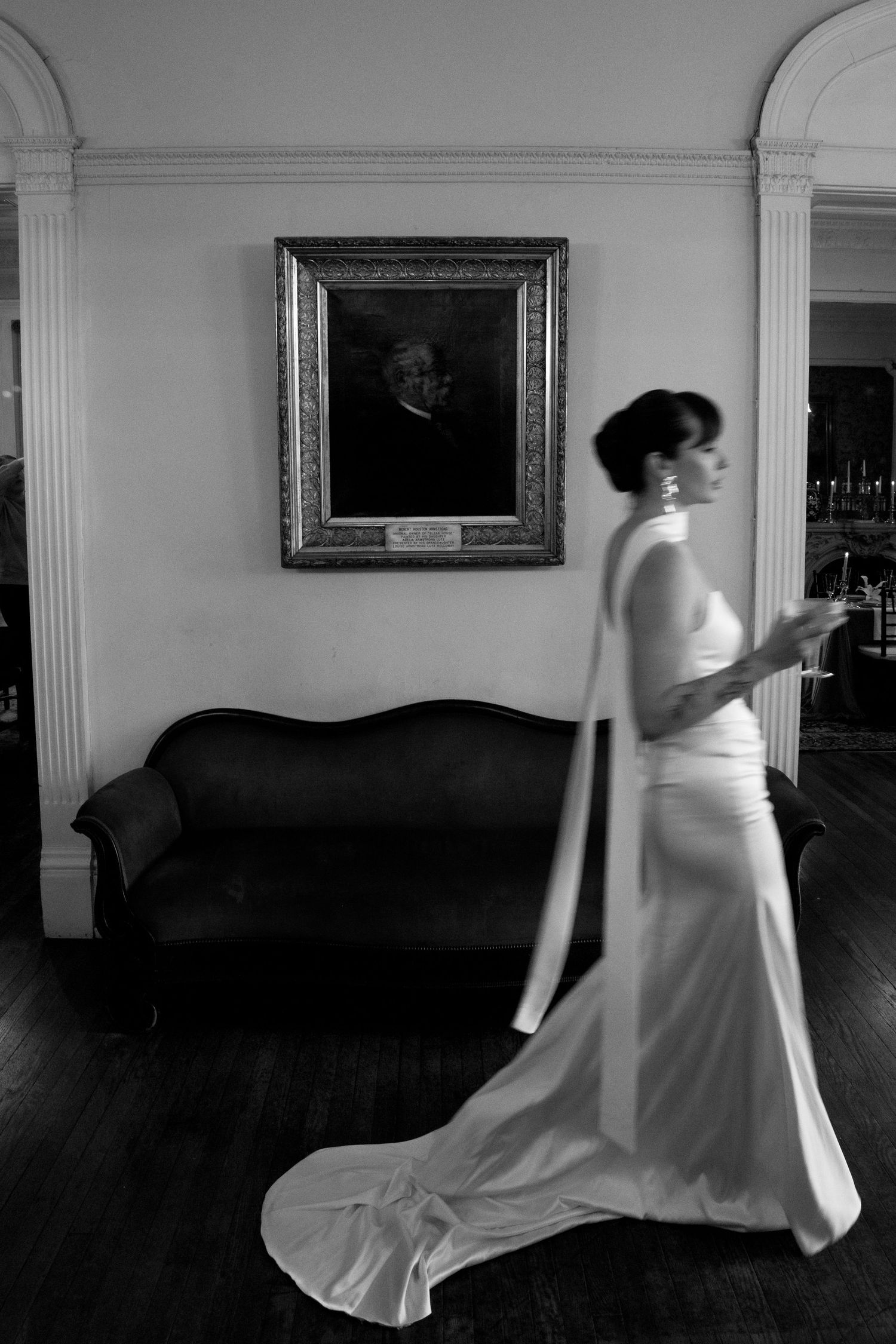 An elegant bride in a flowing white gown walks past a vintage portrait and antique sofa in a black and white photograph.