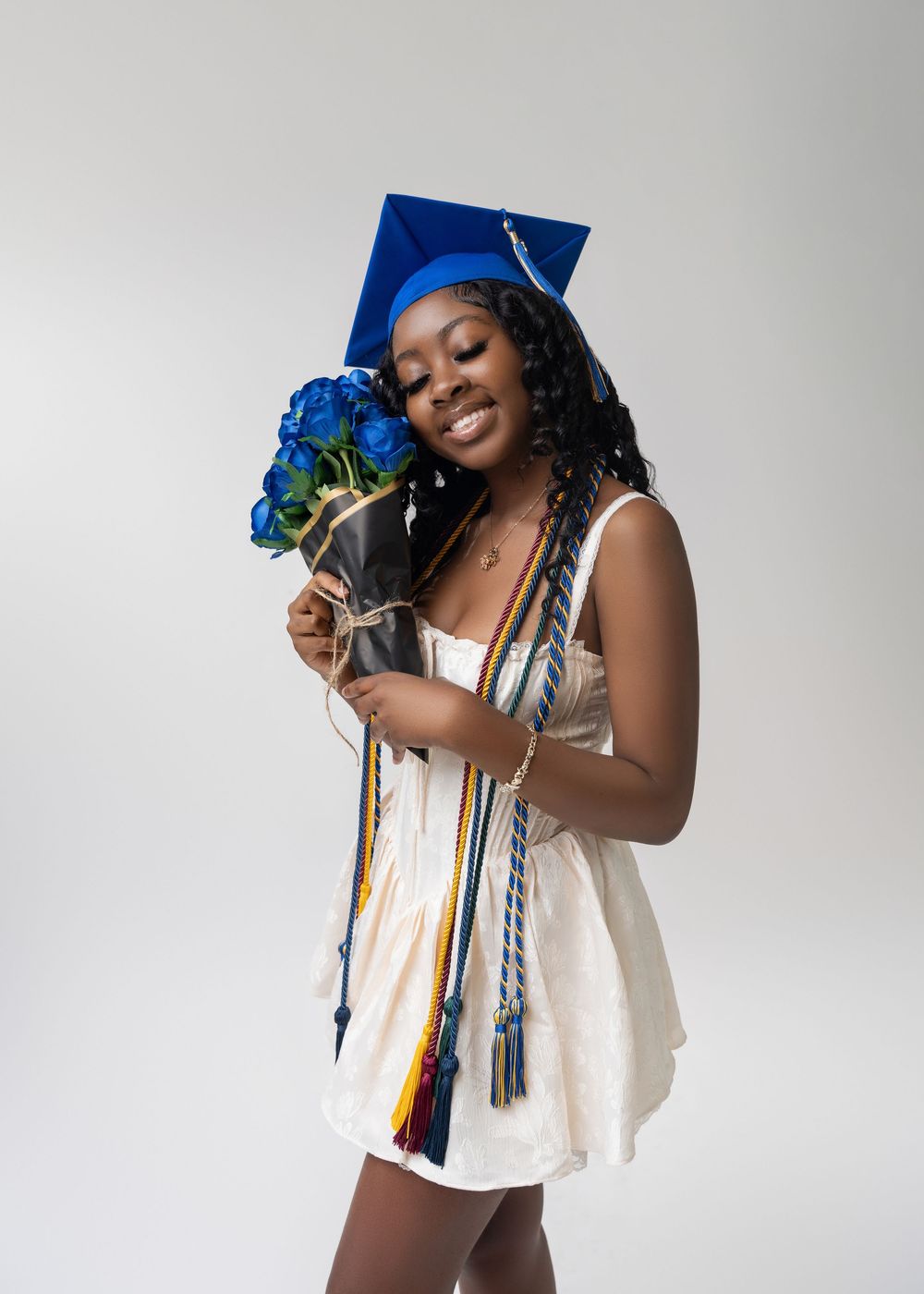 Graduate in white dress holds blue flowers while striking various celebratory poses against light background.