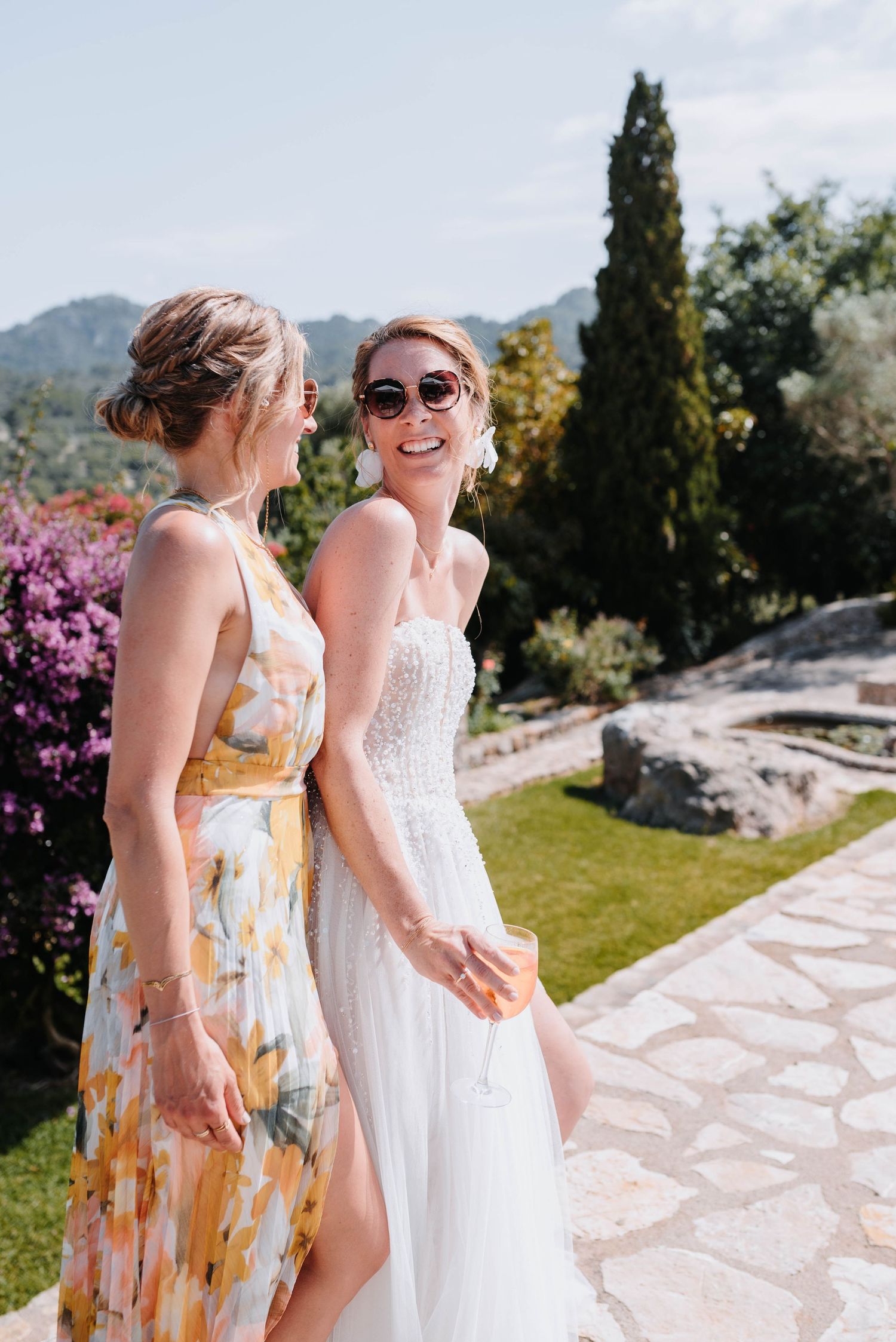 Ein gelbes Blumenkleid bildet einen wunderschönen Kontrast zu einem weißen Hochzeitskleid in einem mediterranen Garten mit Steinmauern.