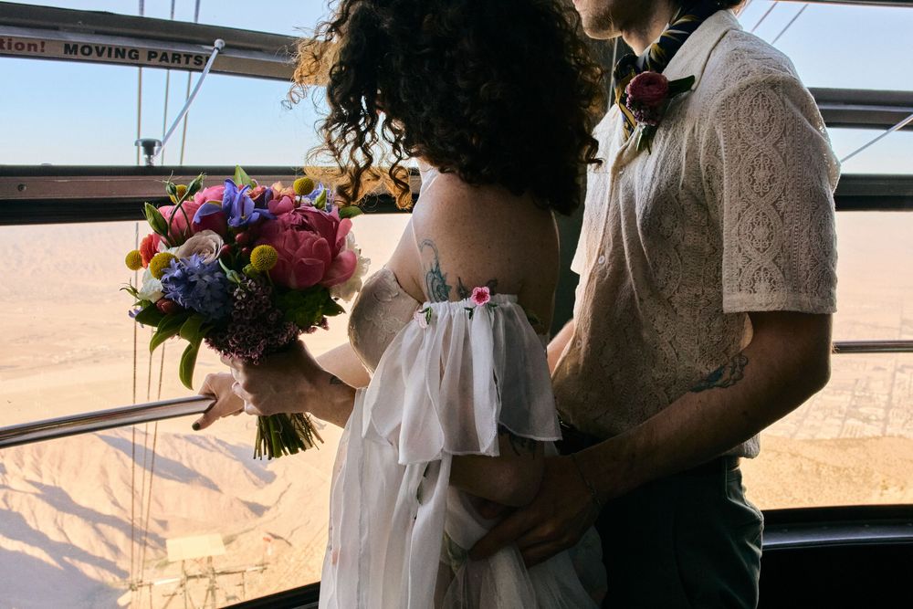 A couple shares an intimate moment during a sunset, with one holding a vibrant bouquet of purple and pink flowers.