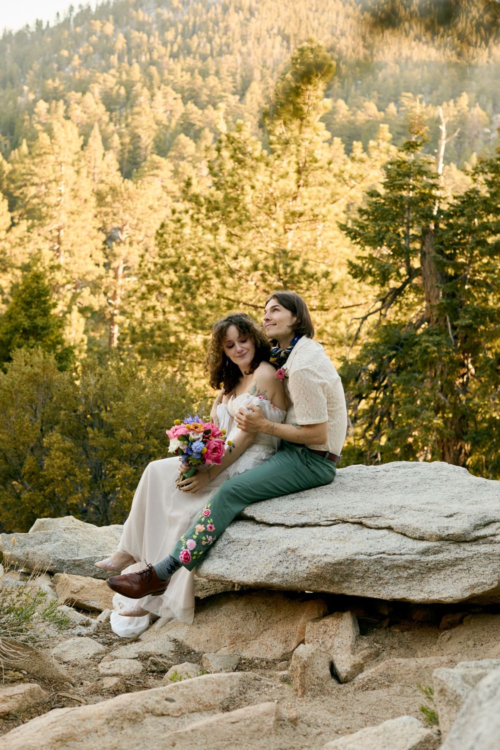 A couple sits together on a large boulder in nature, surrounded by golden autumn trees at sunset.