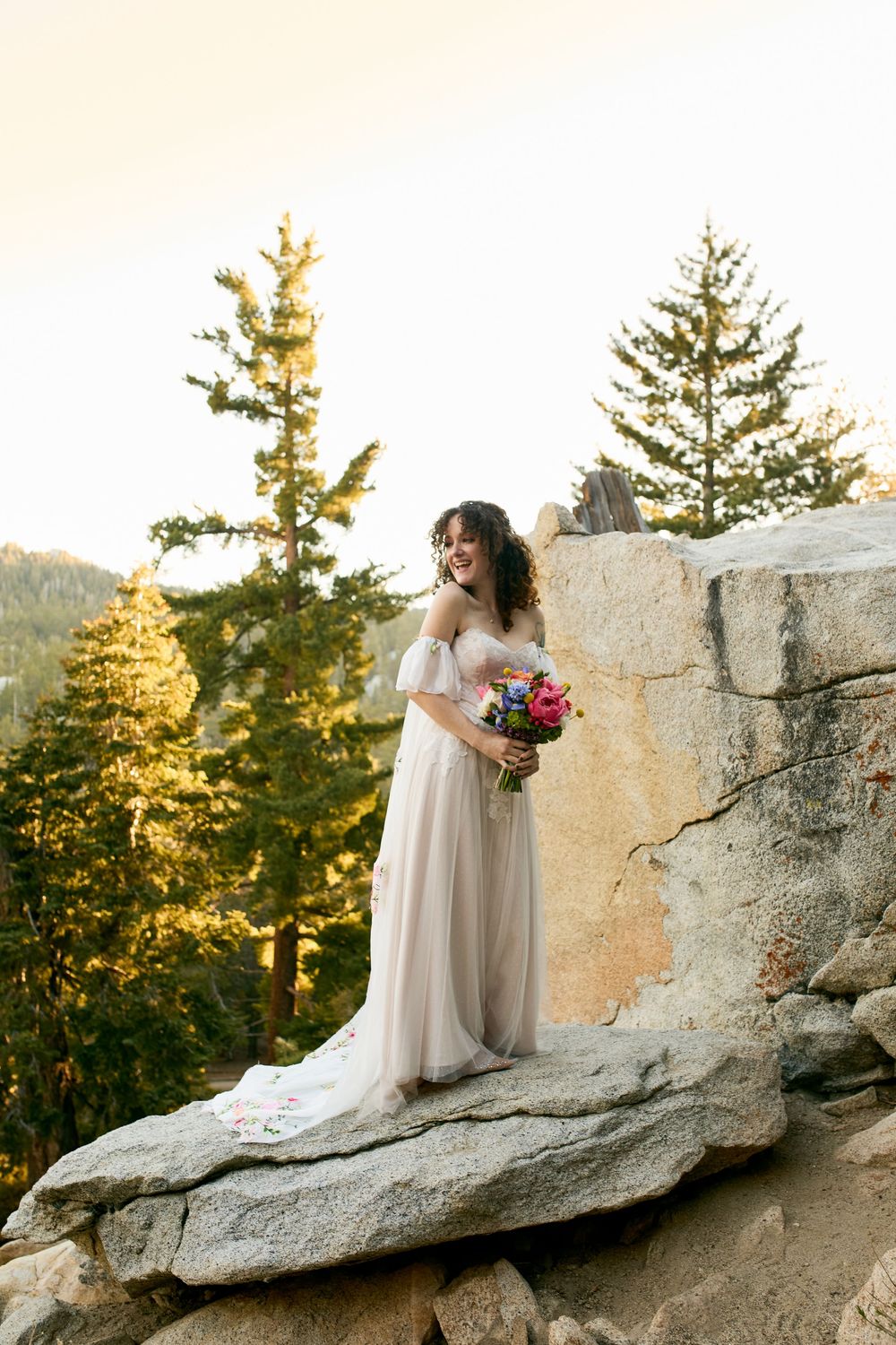 A bride in a flowing white dress stands on a rocky mountain outcrop with evergreen trees and golden sunlight behind her.