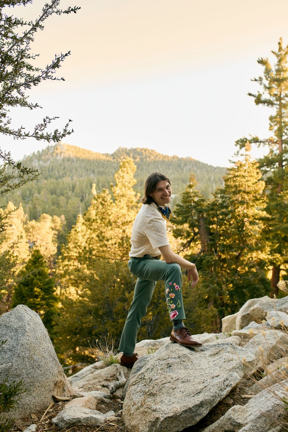 Person climbing and sitting on large granite boulders among evergreen trees during golden sunset in mountainous terrain.