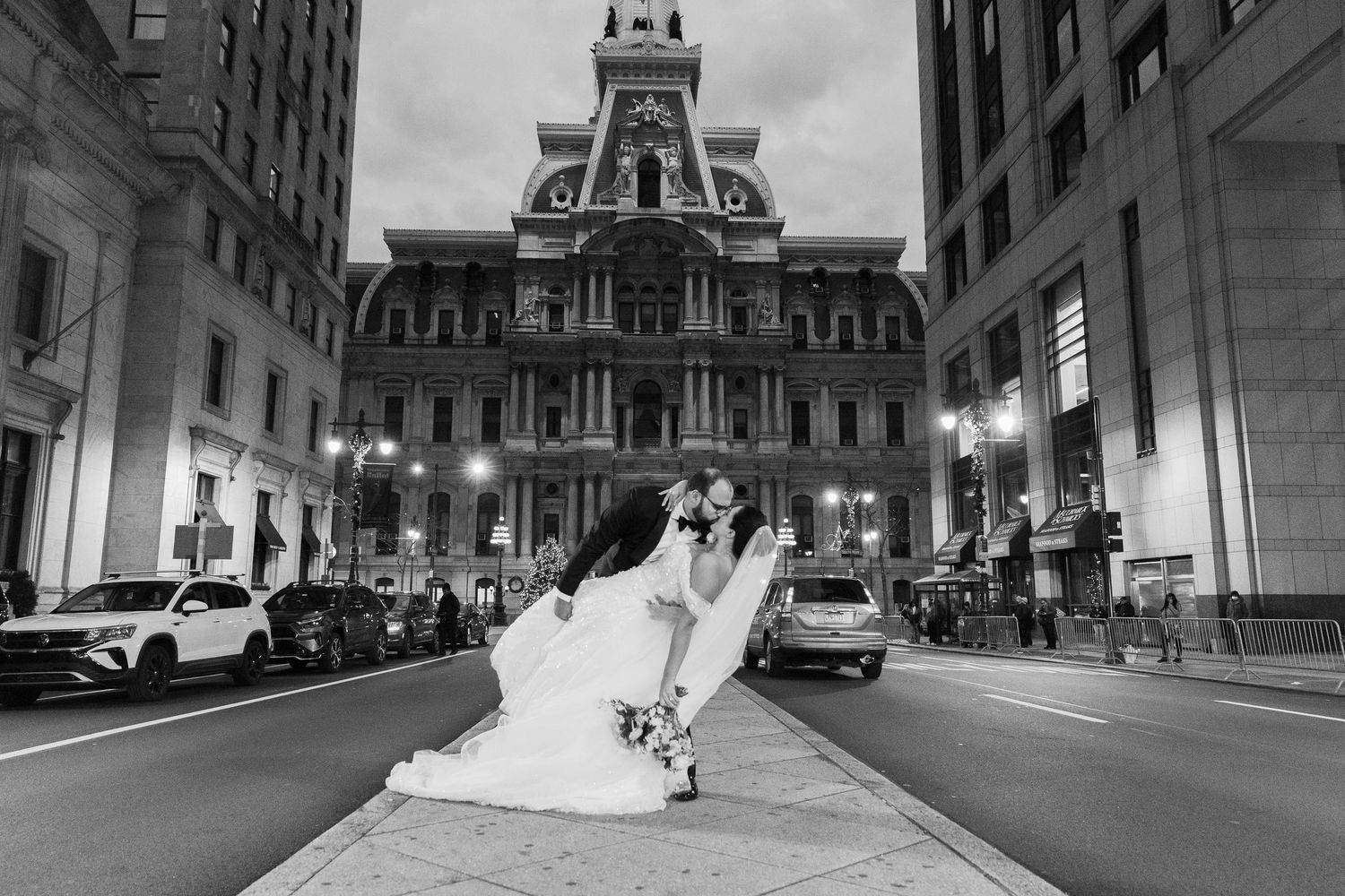 A couple in wedding attire embracing in front of Philadelphia City Hall at dusk with illuminated architecture behind them.