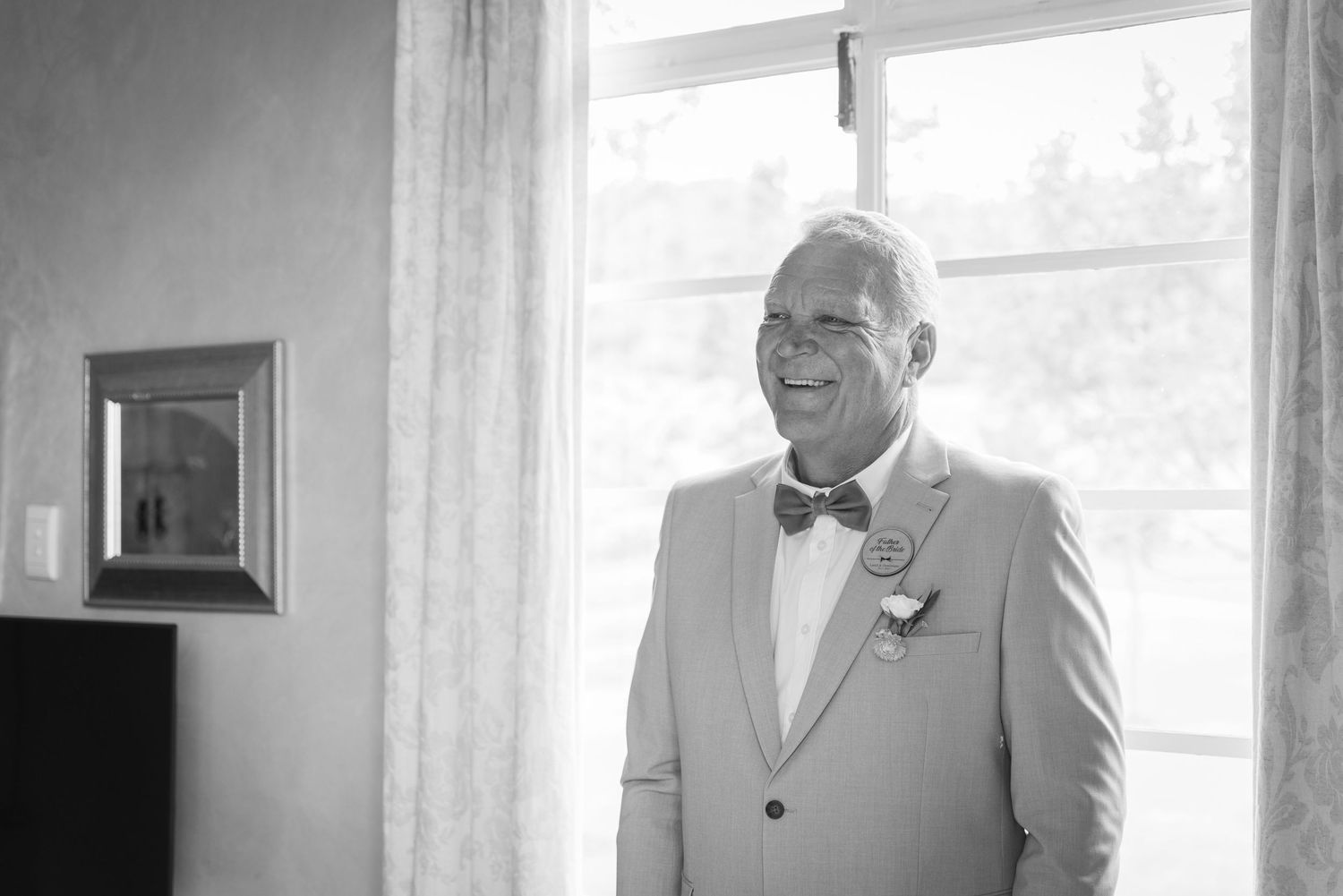 An emotional wedding day moment captured in black and white as groom adjusts his tie by a window.