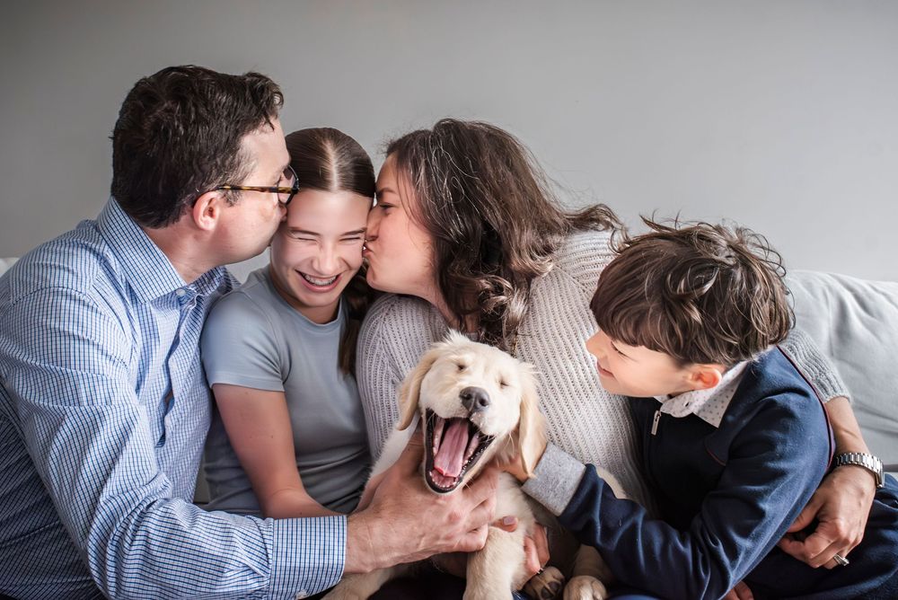 Family of 4 sitting together on the couch and loving on their new puppy, as mom and dad lean in to kiss their oldest child.