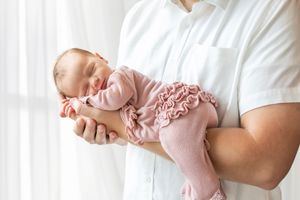 A sleeping newborn baby wearing pink ruffled clothing rests peacefully in someone's arms by a window.