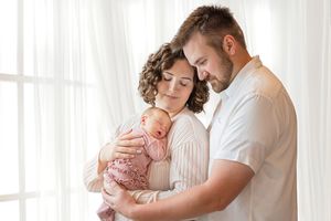 A couple in white embraces their sleeping newborn baby near a bright window in a tender family moment.