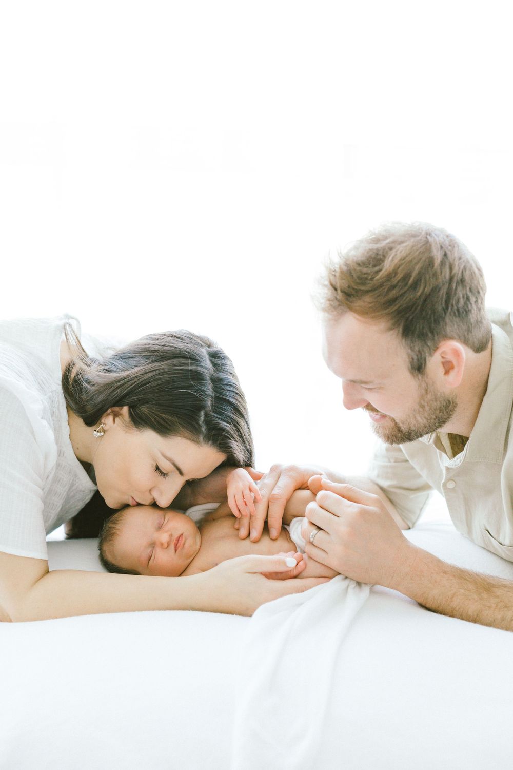 Parents sharing intimate moment with sleeping newborn on white sheets.