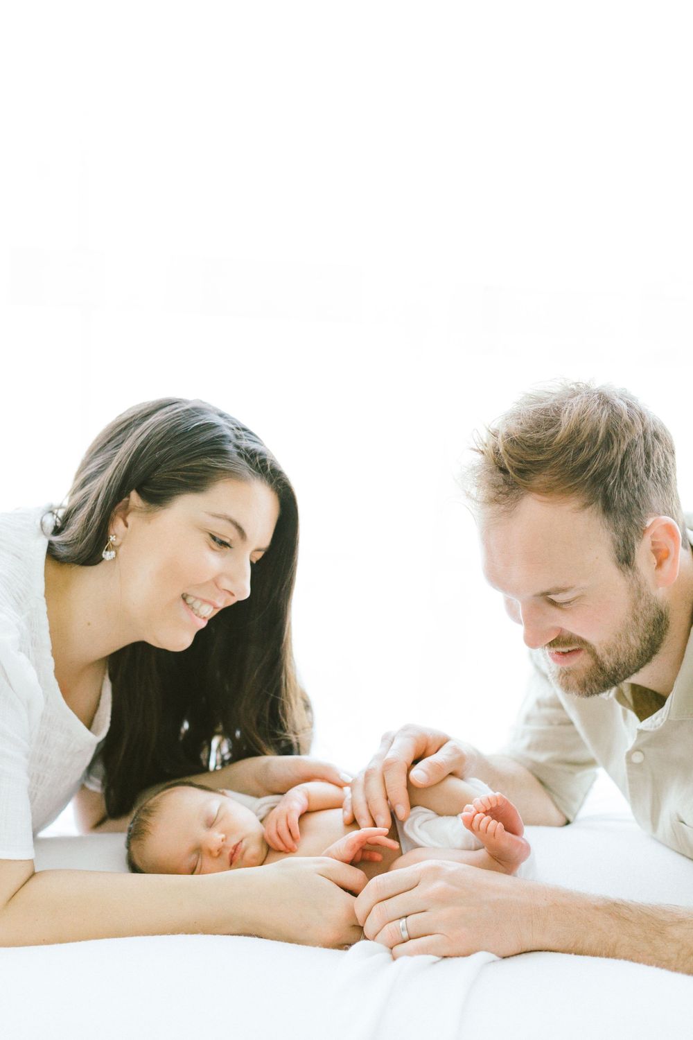 Parents lovingly admiring their newborn baby together on white bedding.