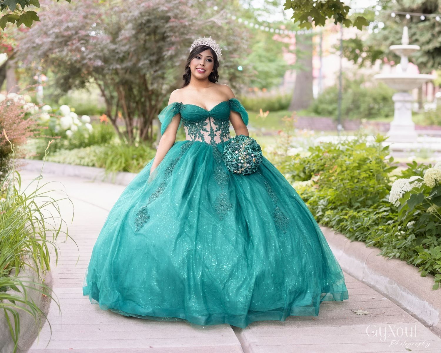 The flowing teal ballgown with off-shoulder neckline creates an elegant silhouette in an outdoor garden setting.