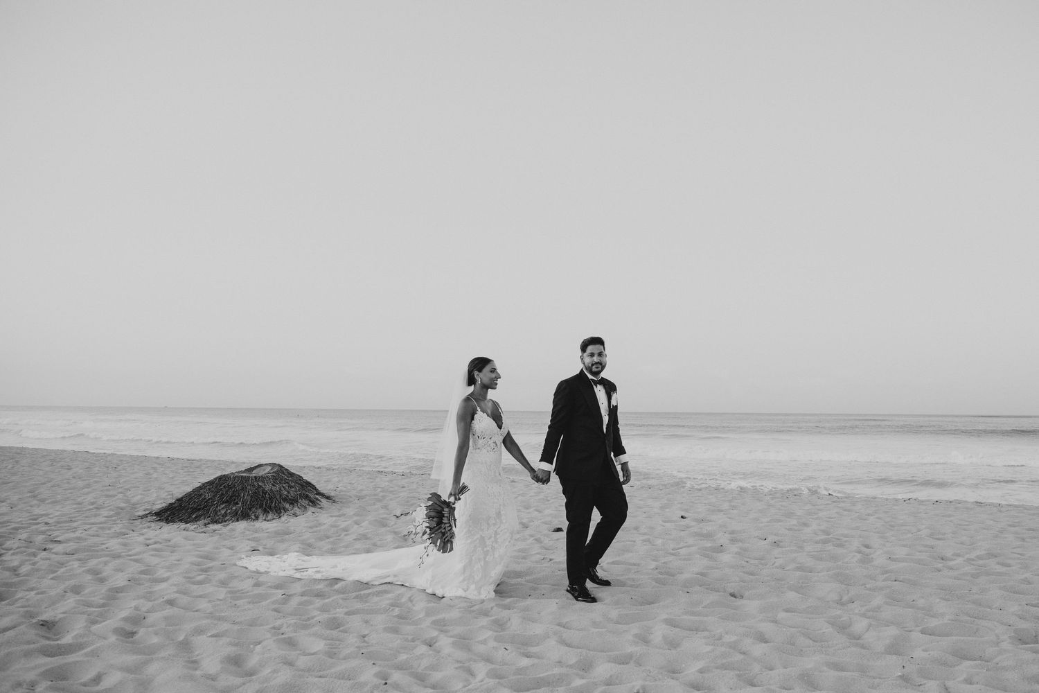 Minimalist black and white photo of newlyweds walking along empty beach shoreline.