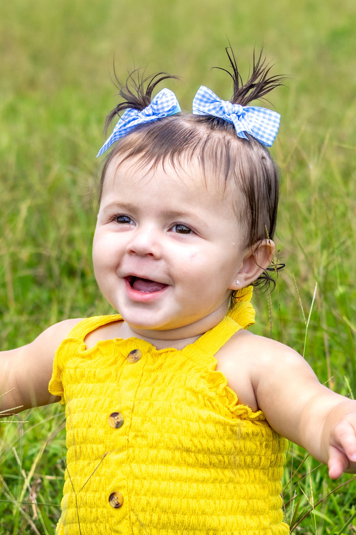 Baby in yellow dress with blue bow smiling and playing in the grass.