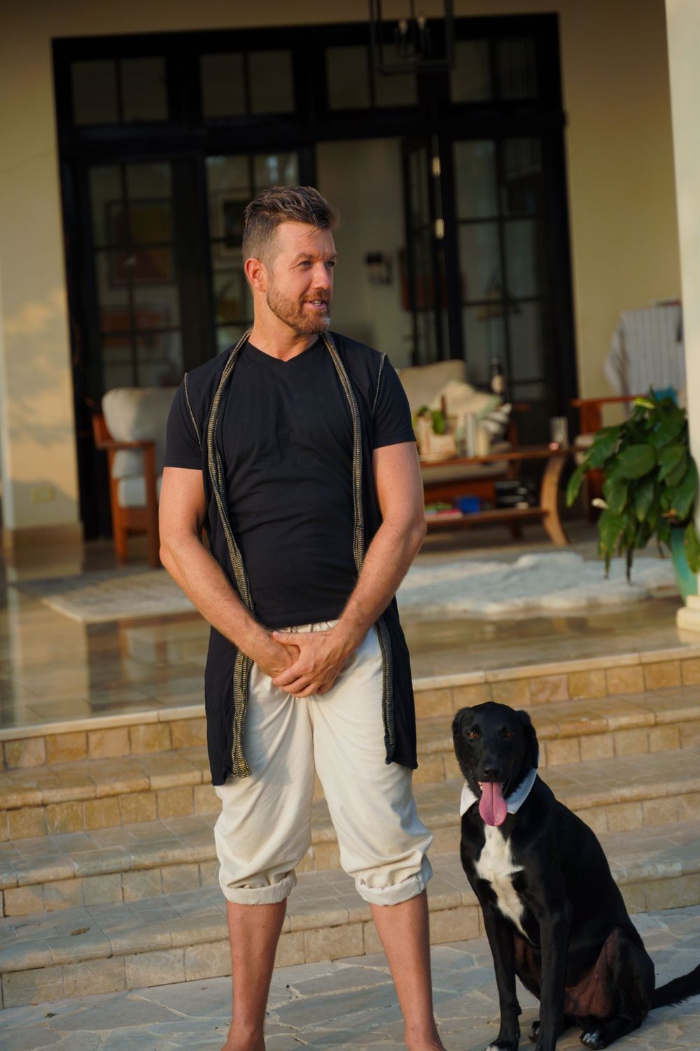 Person wearing black t-shirt and white pants stands on patio steps with a black and white dog by their side.