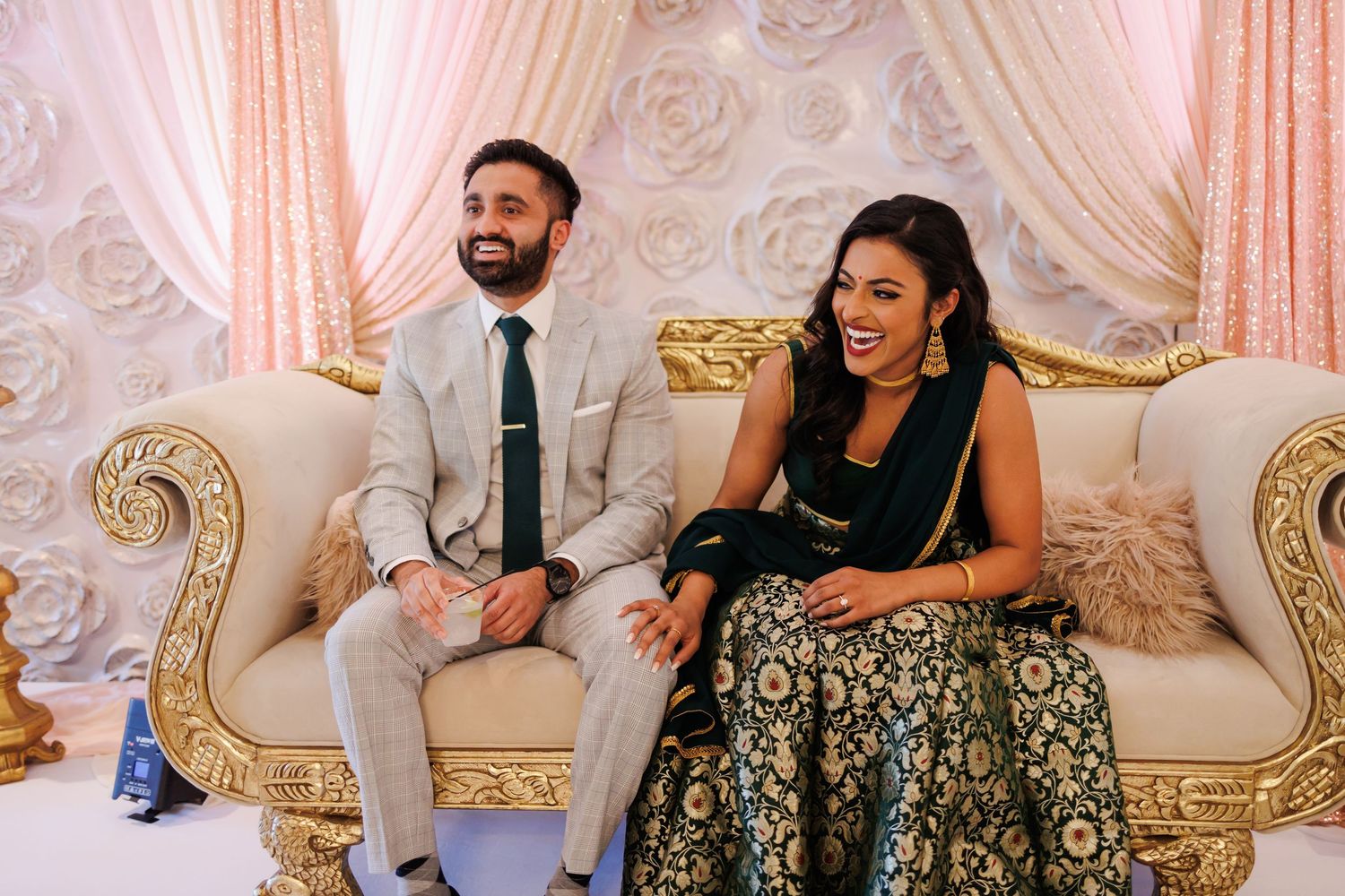 Engaged couple laughing while sitting on an elaborate couch at an Indian betrothal ceremony