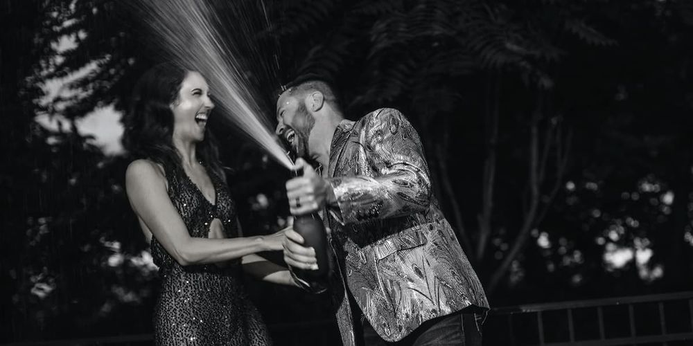 Two people laughing and spraying champagne during a celebration in a dramatic black and white photo.