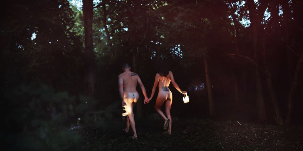 A couple holding hands and walking into dark woods at sunset with one holding a lantern.