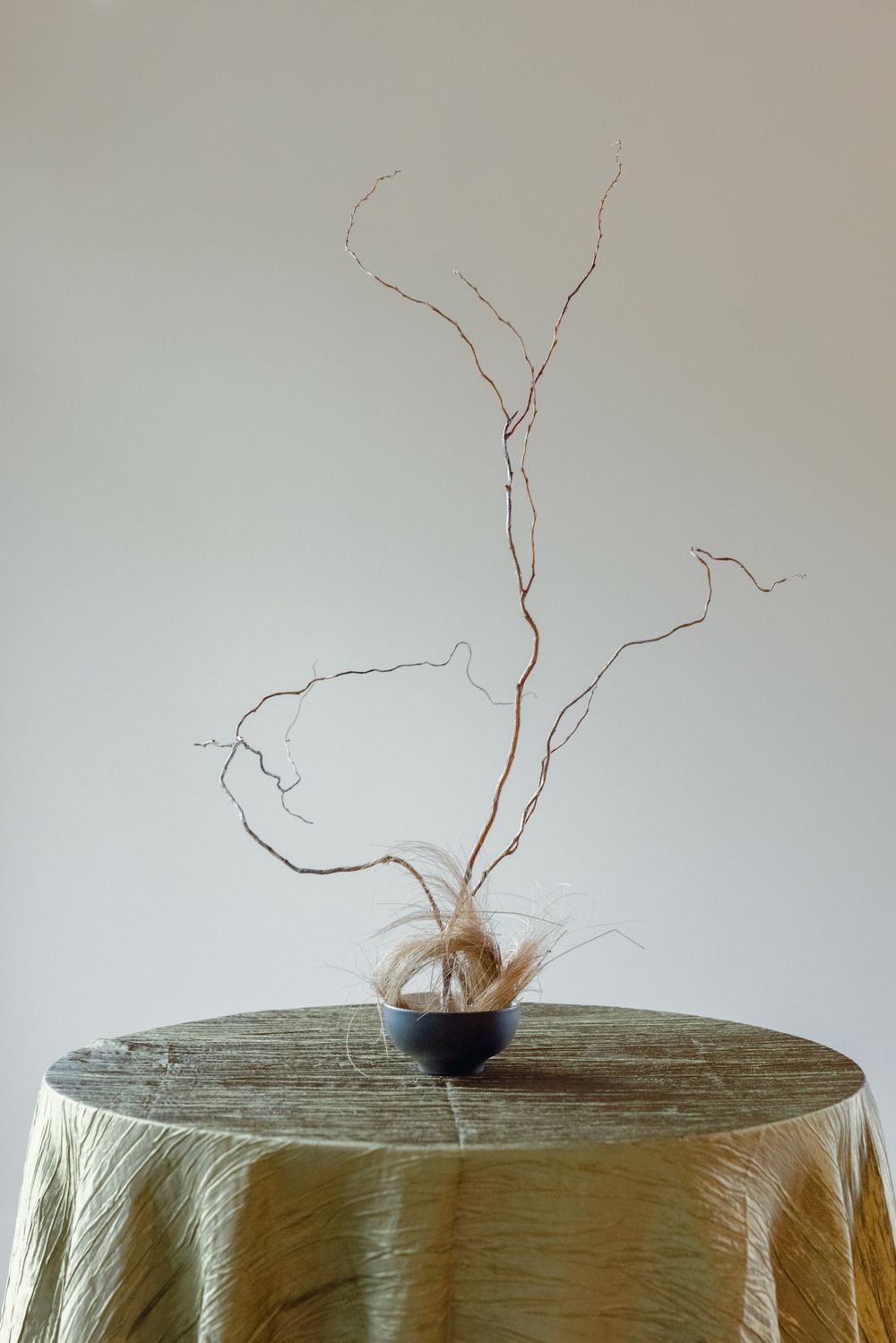 Minimalist dried branch arrangement in dark vessel on textured tablecloth.