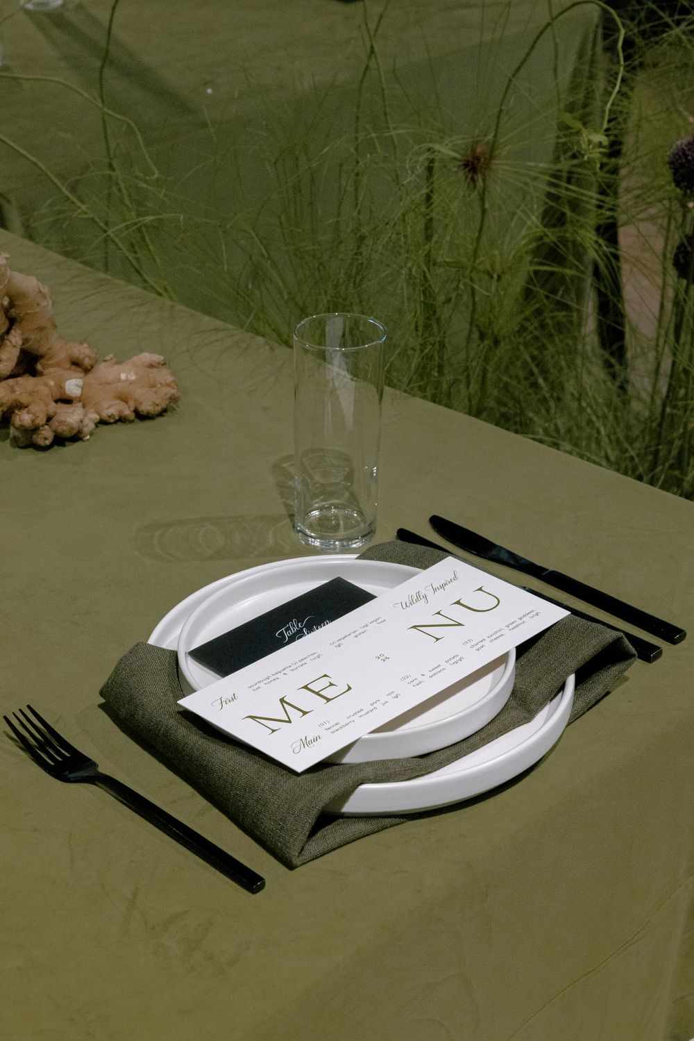 Minimalist table setting with white plates, menu card, and glass against a green backdrop.