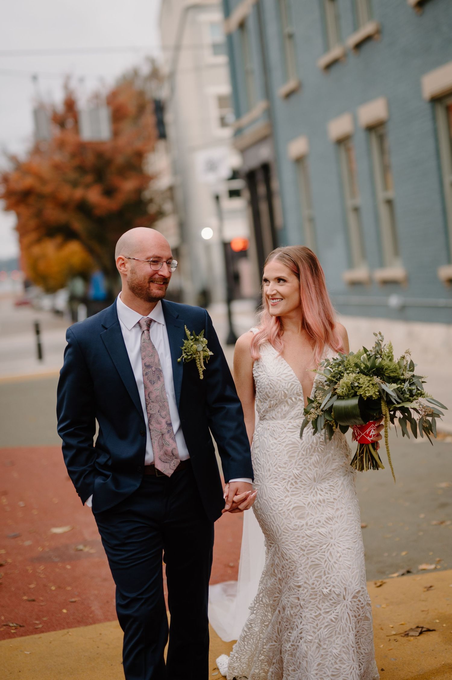 A couple in wedding attire walk together on an autumn day, with the bride holding a green and white bouquet.