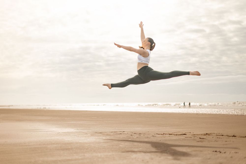 A dancer performs a dramatic split leap against a serene beach landscape at sunset.