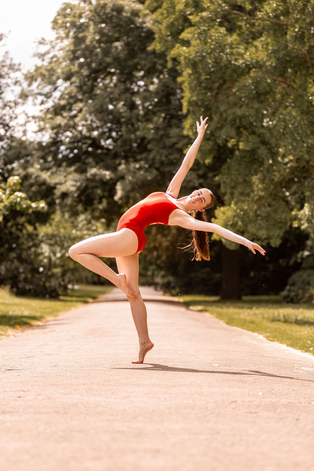 A dancer in a red leotard performs a graceful pose on a sunny path surrounded by green trees.
