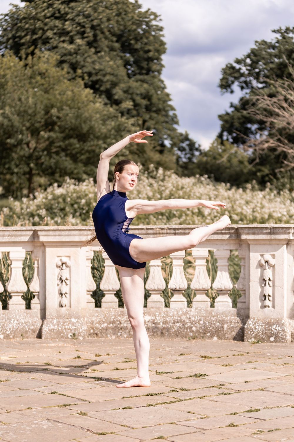 A dancer performs a graceful arabesque sequence on a stone terrace with ornate balustrade and trees in the background.