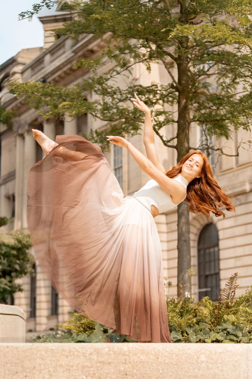 A ballet dancer in a flowing pink dress performs graceful movements in front of a classical building facade.