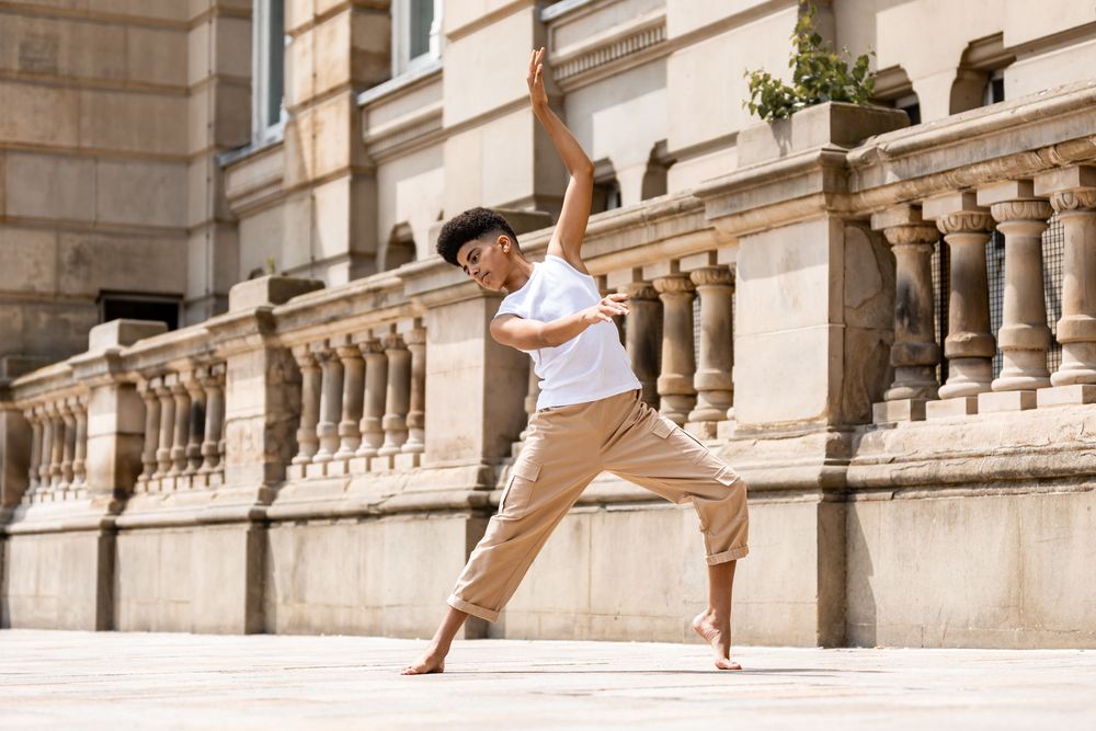 Dancer in casual wear performs graceful leap pose in front of ornate stone balustrade in sunlit urban setting.